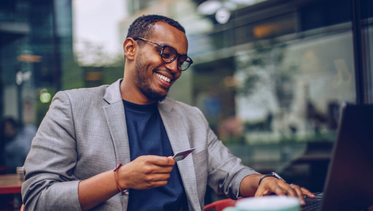 Ein junger, lächelnder Mann sitzt mit einer Kreditkarte in der Hand vor einem Laptop in einem Café.