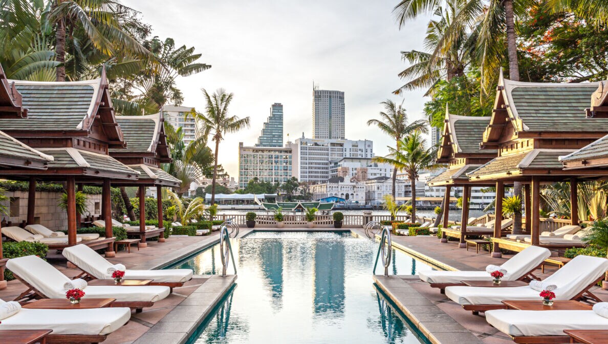 Eine mit Palmen gesäumte Poolanlage im Hotel The Peninsula in Bangkok. Am Wasser stehen Holzliegen mit weißem Bezug, einige befinden sich unter Pagoden-Dächern.
