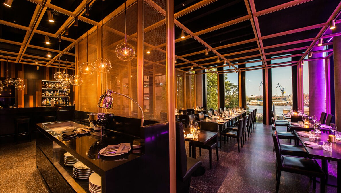 Offener Speiseraum eines stylishen Restaurants mit Blick auf Hafenareal durch Panoramafenster.