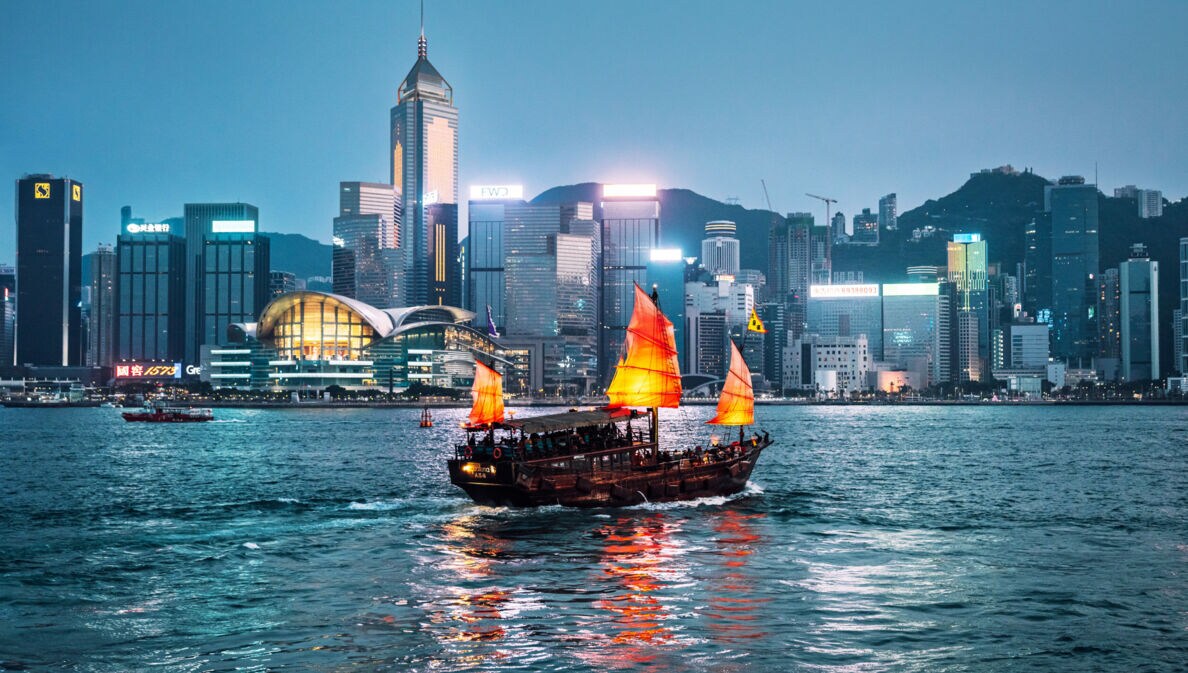 Rote chinesische Dschunke auf dem Wasser vor der Skyline Hongkongs bei Nacht.