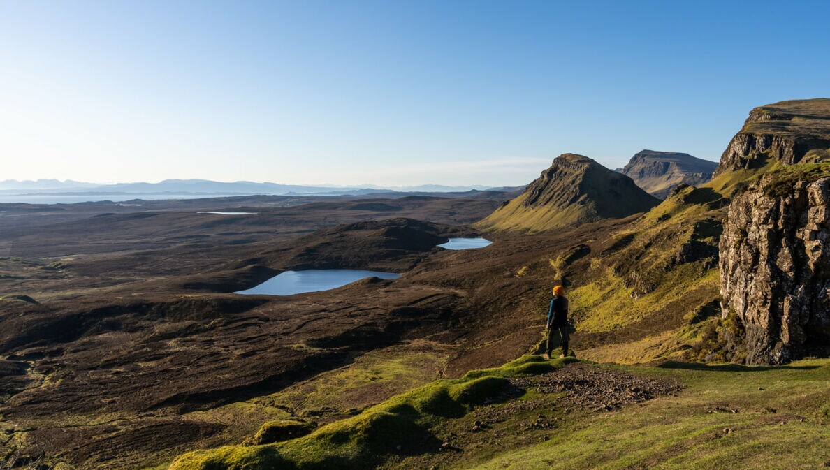 Eine Person steht in einer Landschaft aus Wiesen, Hügeln und Seen in Schottland. 