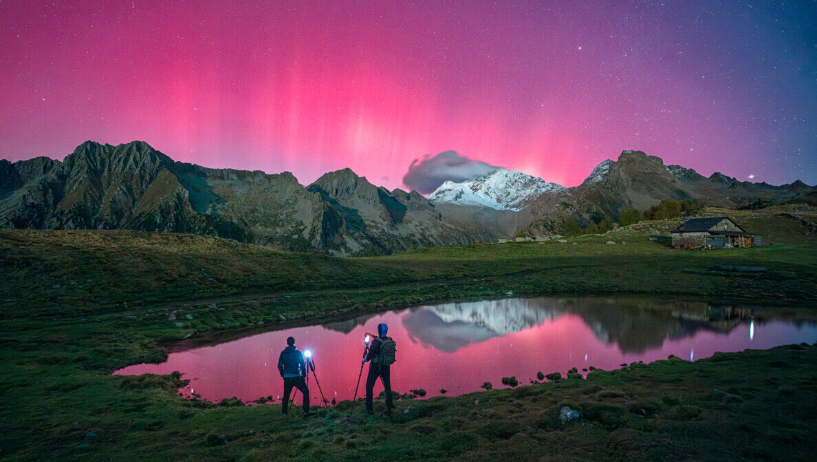 Zwei Menschen stehen vor einer Bergkulisse und fotografieren Polarlichter, die den Himmel pink färben.