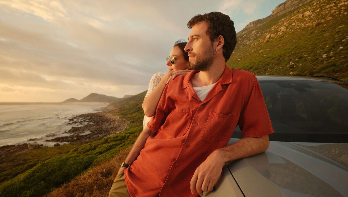Ein Paar lehnt seitlich an einem Auto an einer Küste und blickt aufs Meer.