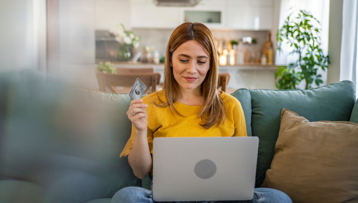 Eine junge Frau im gelben T-Shirt sitzt mit einem Laptop auf dem Schoß auf einem Sofa und schaut lächelnd auf das Display, in ihrer Hand hält sie eine silberne Kreditkarte von American Express.
