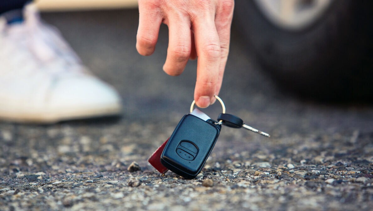 Nahaufnahme einer Hand, die nach einem Autoschlüssel am Asphaltboden greift, dahinter ein Autoreifen und Sneaker in Unschärfe.