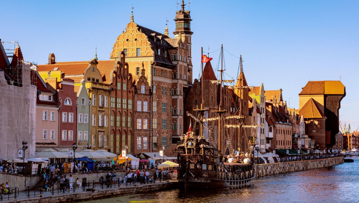 Personen an einer Uferpromenade mit den mittelalterlichen Handelshäusern und historischem Mastschiff.