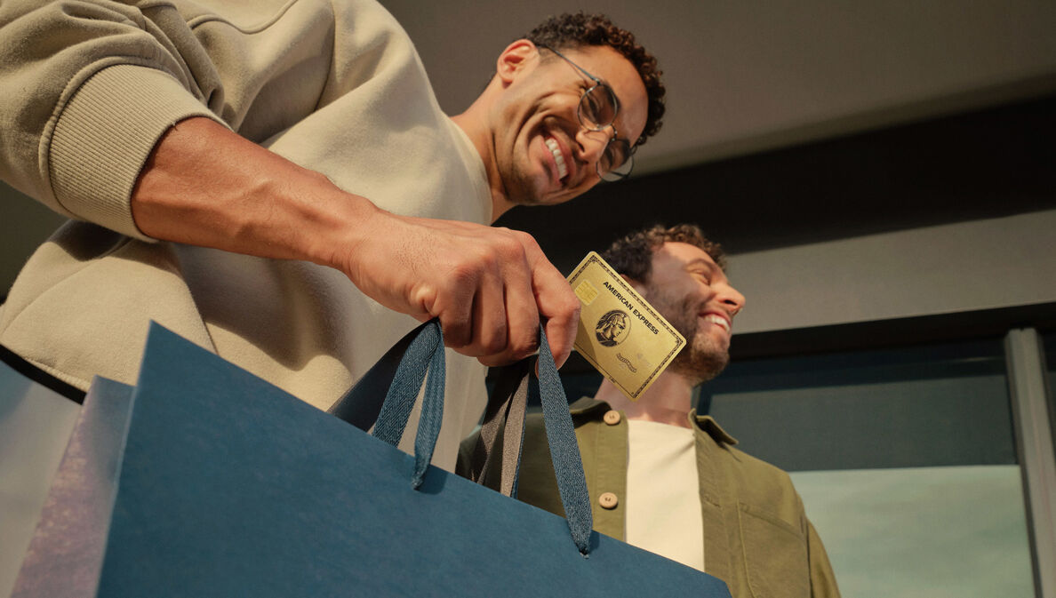 Ein gut gelaunter Mann hält eine goldene Kreditkarte von American Express zusammen mit einer Papiertragetasche in der Hand, daneben ein weiterer Mann.