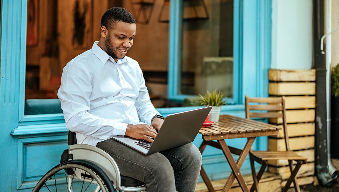 Ein junger Mann im Rollstuhl arbeitet am Laptop auf der Terrasse eines Cafés.