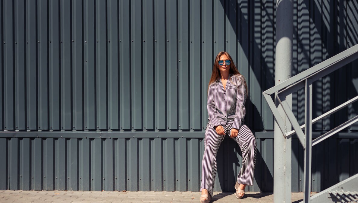 Junge Frau mit Sonnenbrille in blau-weiß gestreiftem Pyjama lehnt an eine Wand aus grauem festem Wellblech.