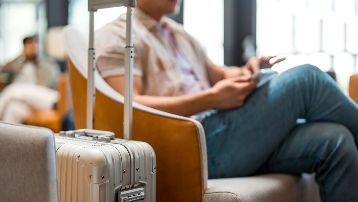 Ein Mann sitzt im Wartebereich eines Flughafens. Im Vordergrund steht sein Rollkoffer.