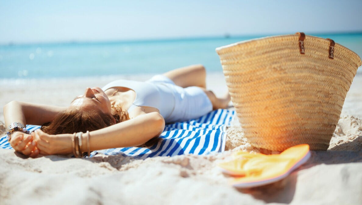 Eine Frau liegt rücklings entspannt auf einem gestreiften Badehandtuch an einem Sandstrand am Meer, neben ihr ein Strohkorb und Badeschuhe.