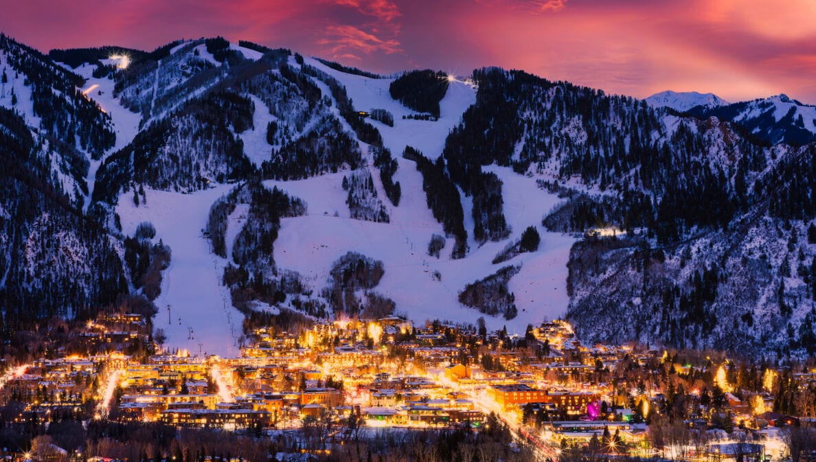 Bergdorf mit beleuchteten Gebäuden und schneebedeckten Skipisten bei Sonnenuntergang.