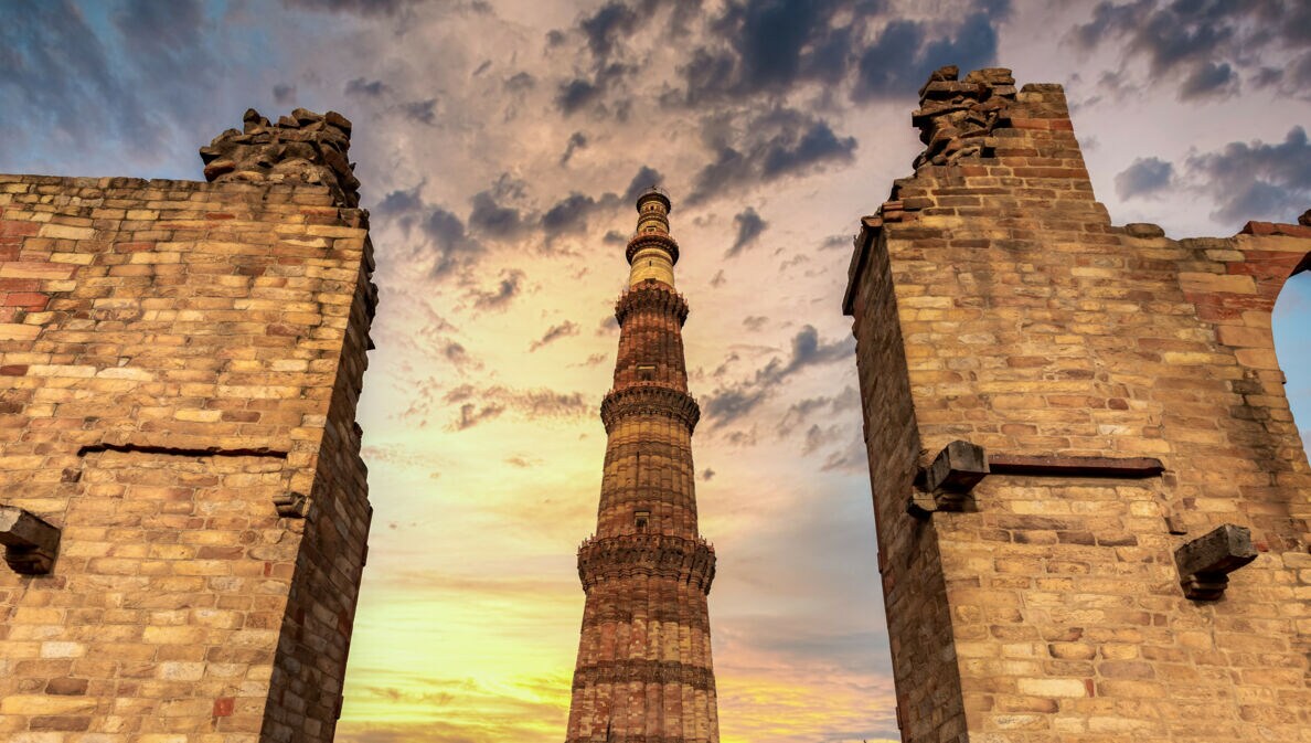 Das Qutub Minar, ein hoher roter Sandsteinturm, umgeben von Ruinenmauern bei Sonnenuntergang mit bewölktem Himmel