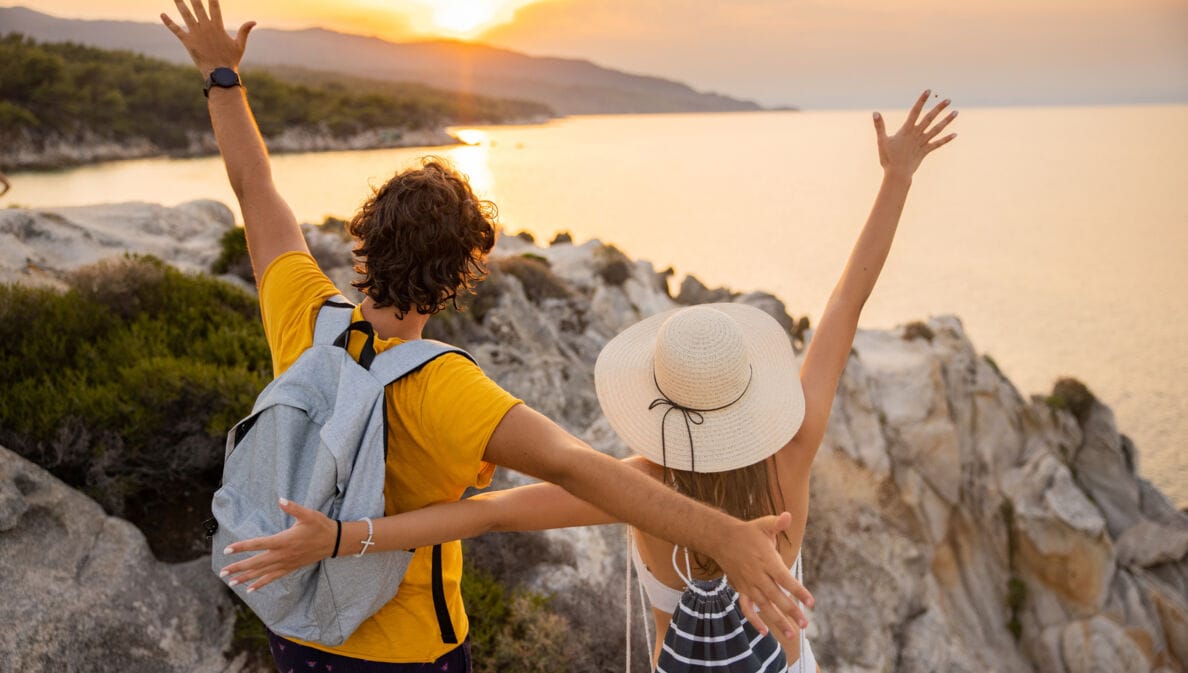 Ein Paar steht mit offenen Armen auf einem Felsen und schaut auf das Meer und den Sonnenuntergang.
