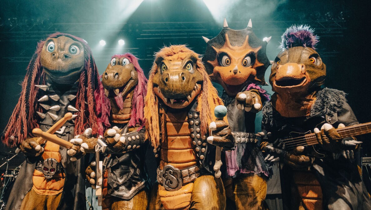 Vier Personen in Dinosaurierkostümen, die in die Kamera gucken und teilweise Instrumente und ein Mikrofon halten
