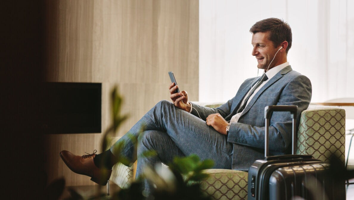Ein Geschäftsmann sitzt in einem Sessel in einer Flughafen-Lounge und schaut lächelnd auf sein Smartphone.