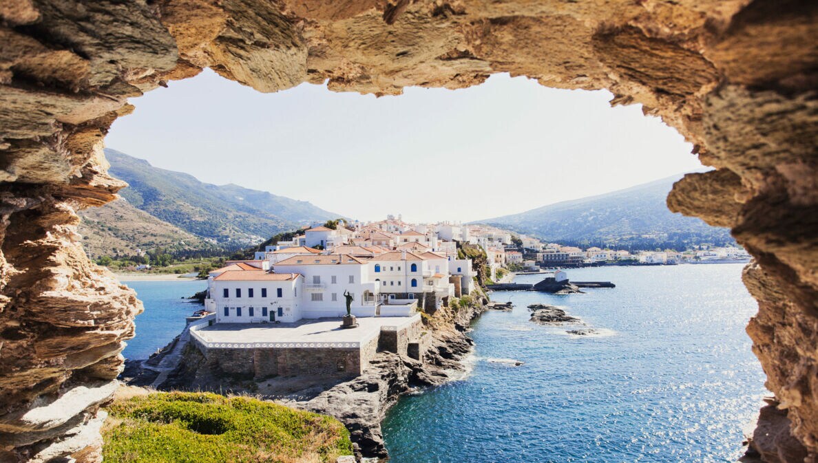 Blick auf weiße Häuser eines Ortes auf einer felsigen Landzunge im Meer durch ein Loch in einer Felsenmauer.