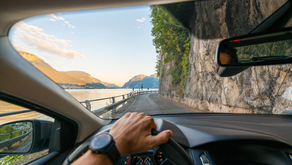 Blick auf eine Küstenstraße entlang einer Felswand durch die Windschutzscheibe eines Autos, im Vordergrund eine Hand am Lenkrad.