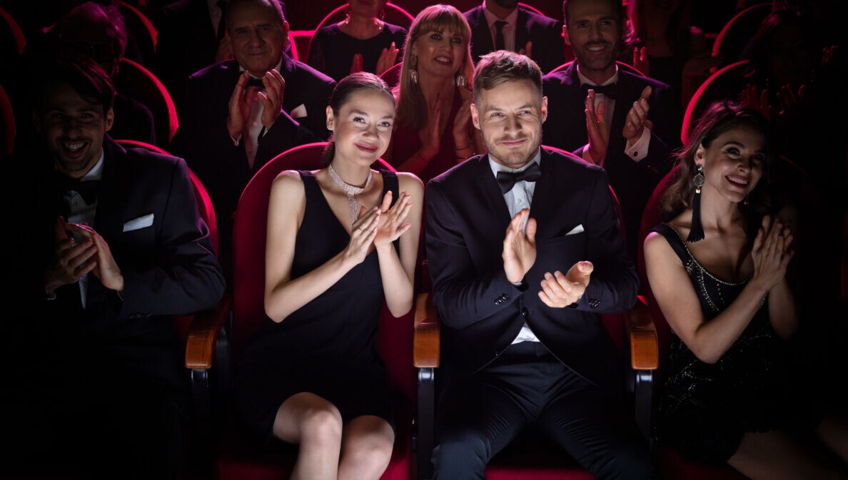 Zwei in Schwarz gekleidete Personen sitzen klatschend auf roten Sesseln im Publikum.