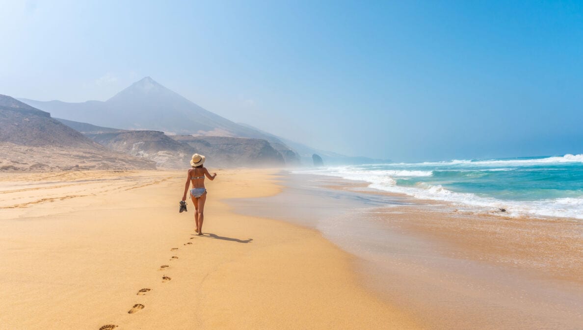 Rückansicht einer Frau im Bikini, die einen breiten Sandstrand entlangläuft, im Hintergrund spitze Felsformationen.