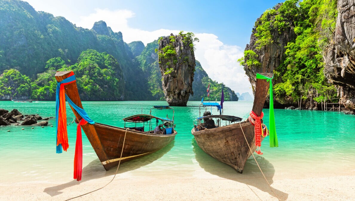 Zwei Holzboote an einem Sandstrand in einer Meeresbucht mit türkisem Wasser, umgeben von begrünten Felsen.