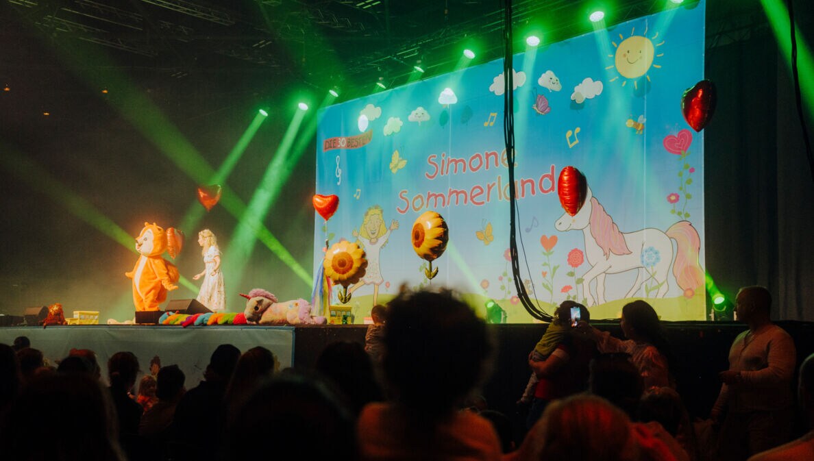 Simone Sommerland steht auf einer Bühne voller Luftballons, neben ihr eine Person im Eichhörnchen-Kostüm, vor ihr Publikum.