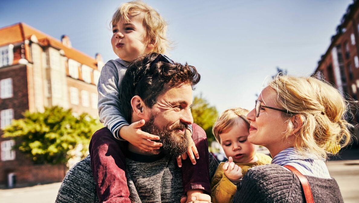 Vater trägt lachendes Kleinkind auf Schultern, Mutter mit Brille schaut liebevoll, während sie ein weiteres Kleinkind im Arm hat.