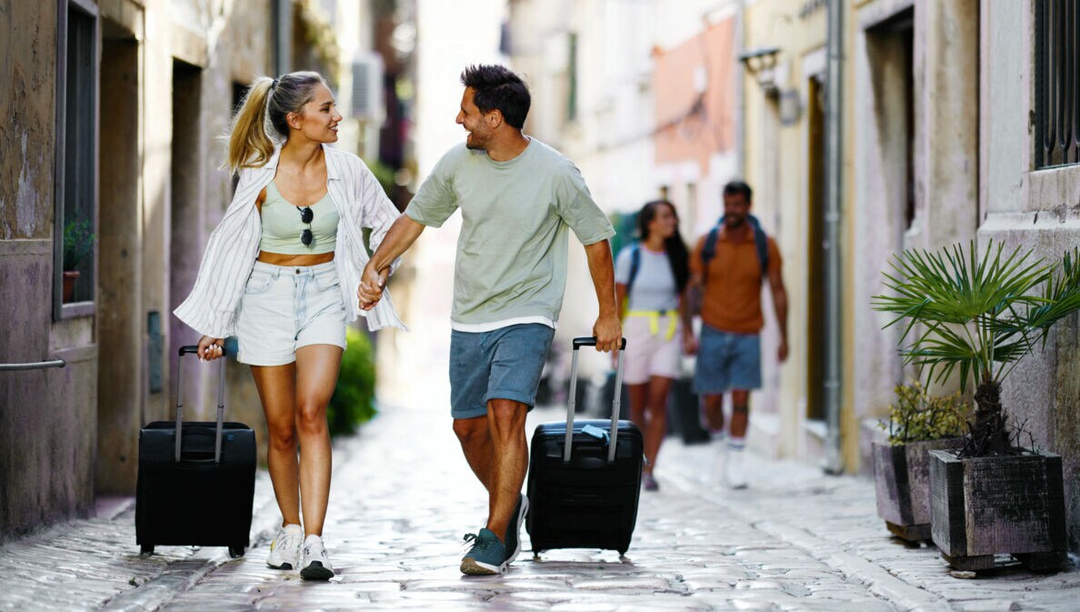 Ein händchenhaltendes Paar in Sommerkleidung geht mit Rollkoffern durch eine Gasse mit Kopfsteinpflaster.