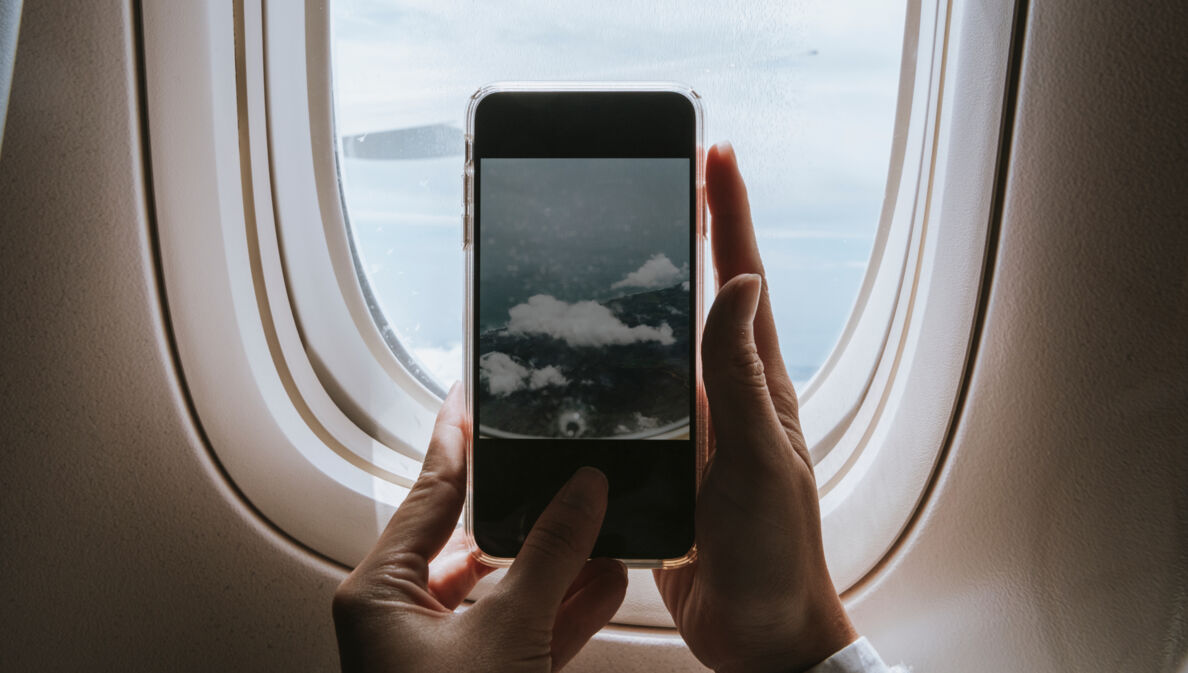Hände halten ein Smartphone vor einem Flugzeugfenster, das den Himmel mit Wolken zeigt.