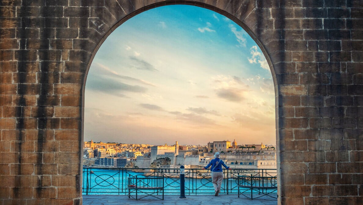 Blick durch einen großen Steinbogen auf eine Stadt am Wasser bei Sonnenuntergang, eine Person lehnt am Geländer