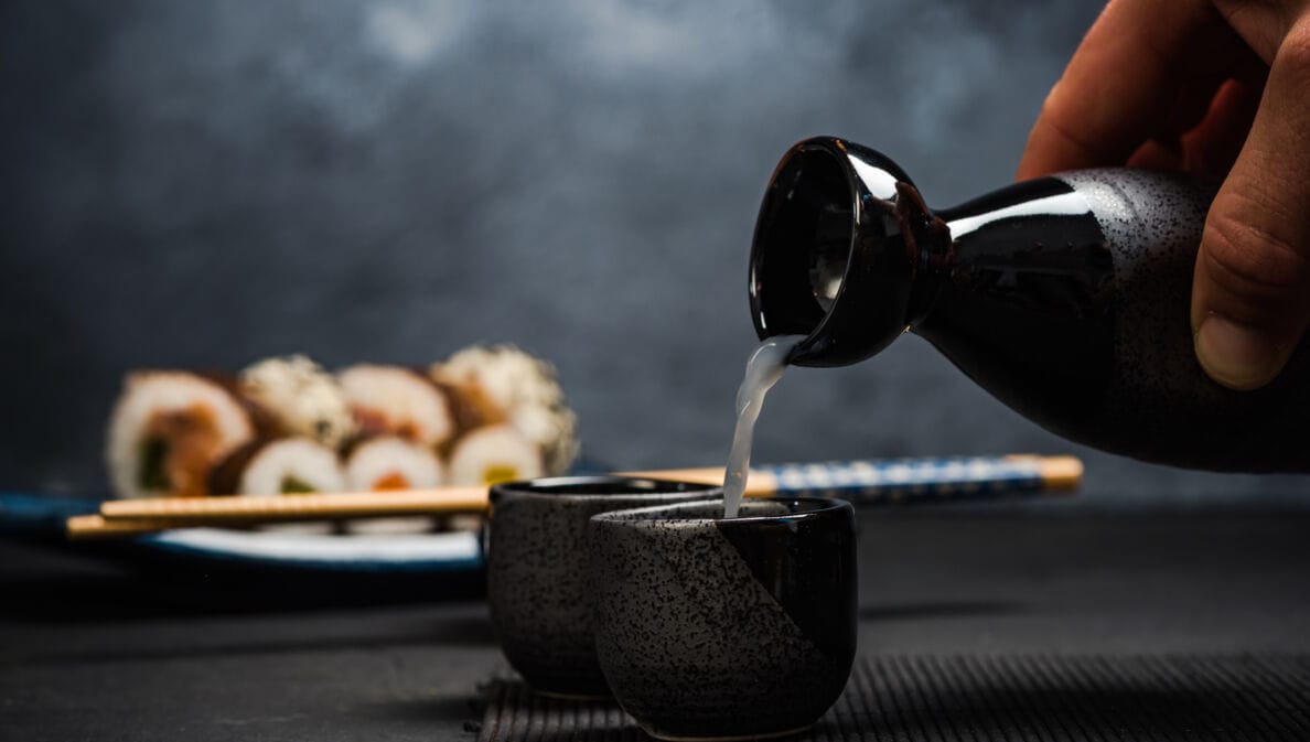 Hand gießt Sake aus einer schwarzen Karaffe in eine kleine schwarze Tasse, im Hintergrund Sushi und Essstäbchen