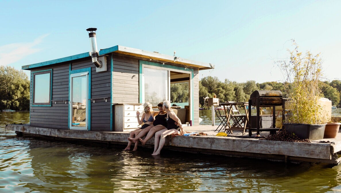 Drei Frauen in Badekleidung sitzen am Rand eines Pontons mit Hausboot und lassen ihre Füße ins Wasser baumeln bei Sonnenschein.