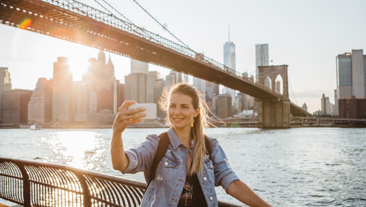 Frau macht Selfie vor der Brooklyn Bridge und der Skyline von New York bei Sonnenuntergang