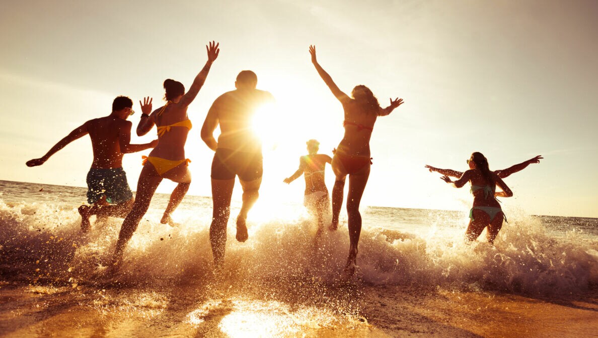 Gruppe von sechs jungen Menschen läuft im Sonnenuntergang ins Meer, Wasser spritzt um sie herum.