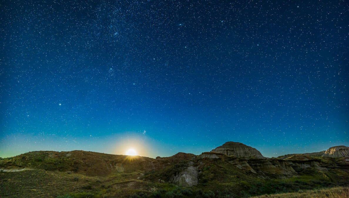 Klarer, tiefblauer Sternenhimmel mit aufgehendem Mond über einer felsigen Graslandschaft.