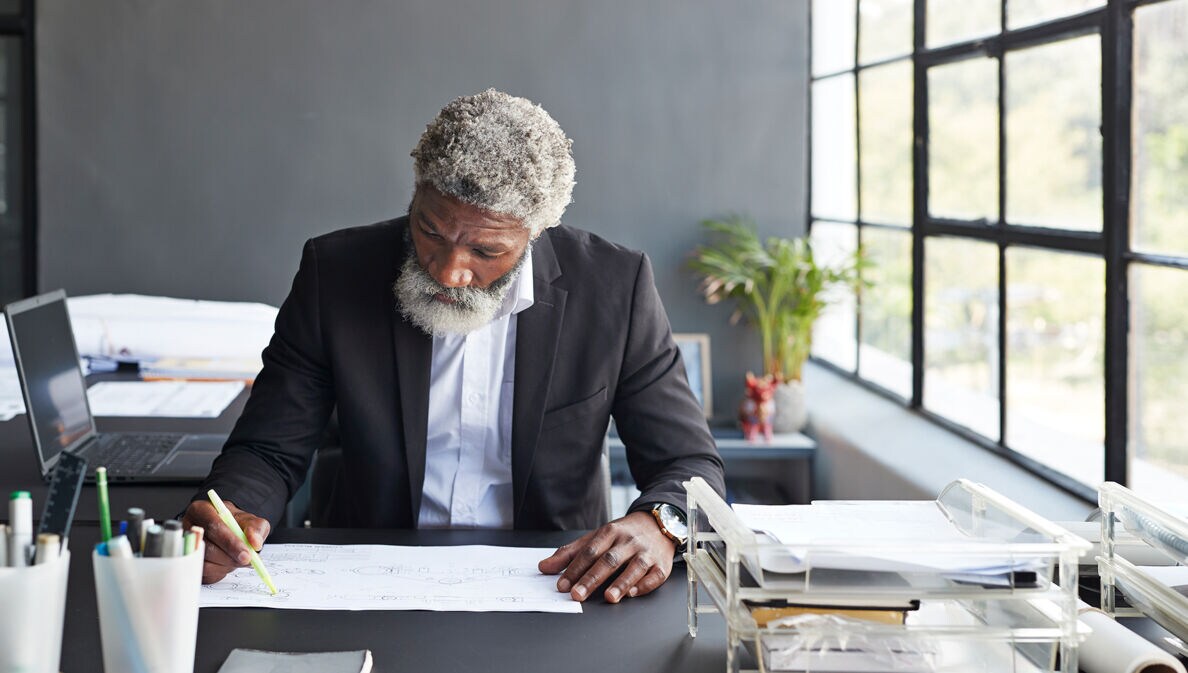 Ein älterer Mann arbeitet fokussiert im Büro an einer technischen Zeichnung.