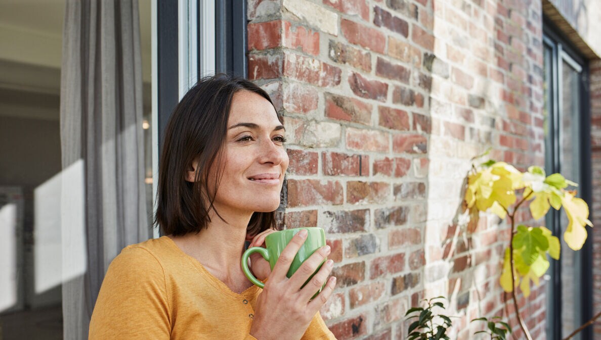 Eine Frau mit einer Tasse in der Hand lehnt entspannt an der Hauswand.