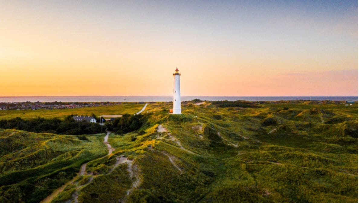 Weißer Leuchtturm auf grünen Dünen bei Sonnenuntergang mit Dorf und Meer im Hintergrund.