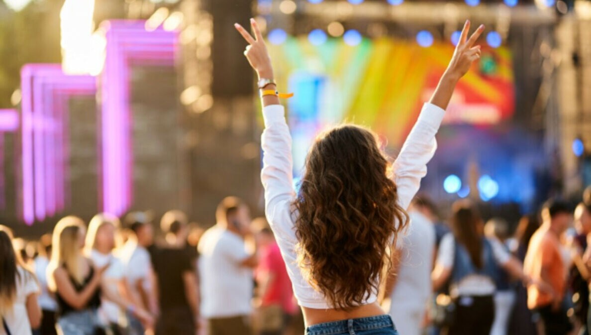 Rückansicht einer jungen Frau, die ihr Hände mit Peace-Zeichen in die Luft hebt vor einer Musikbühne mit Publikum im Freien.