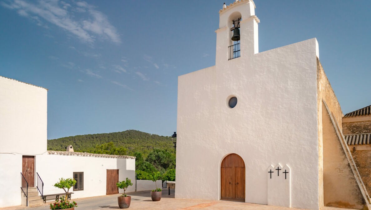 Weiße Kirche mit Glockenturm und drei Kreuzen an der Wand, umgeben von Gebäuden und Bergen unter blauem Himmel