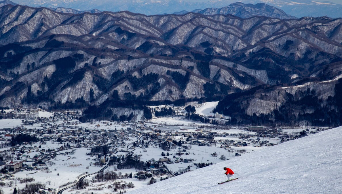 Skifahrer in roter Jacke fährt einen schneebedeckten Hang hinab mit Blick auf ein verschneites Dorf und bewaldete Berge im Hintergrund