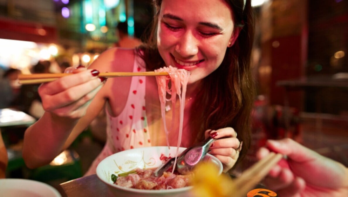 Nahaufnahme einer jungen, lächelnden Frau beim Essen eines asiatischen Nudelgerichtes mit Essstäbchen auf einem Straßenmarkt bei Nacht.