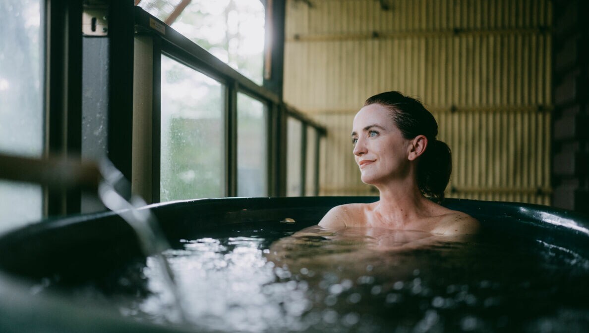 Eine Frau sitzt in einem Thermalbad aus dunklem Stein in einem japanischen Spa mit Bambuswand und schaut zufrieden durch ein Fenster hinaus.
