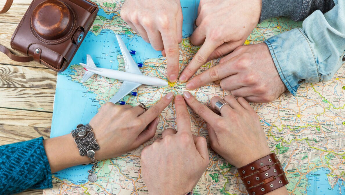 Sechs Personen, von denen nur die Hände zu sehen sind, zeigen auf einen Punkt auf einer Landkarte. Daneben liegen ein alter Fotoapparat und ein Modell eines Flugzeugs.