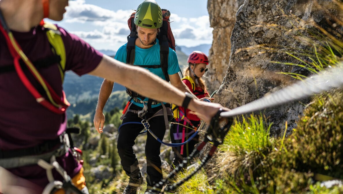 Drei Personen in Kletterausrüstung an einem Felsen mit Drahtseil.