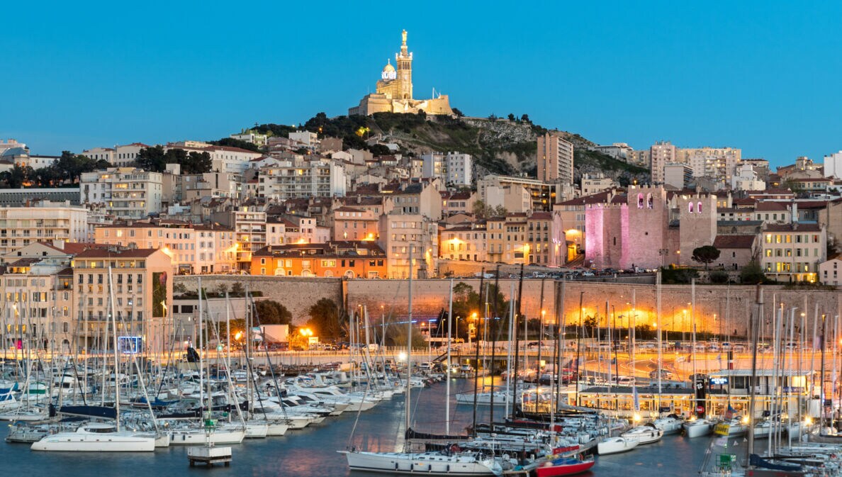Abendliche Hafenansicht von Marseille mit Segelbooten im Wasser und der beleuchteten Kirche Notre-Dame de la Garde auf einem Hügel im Hintergrund