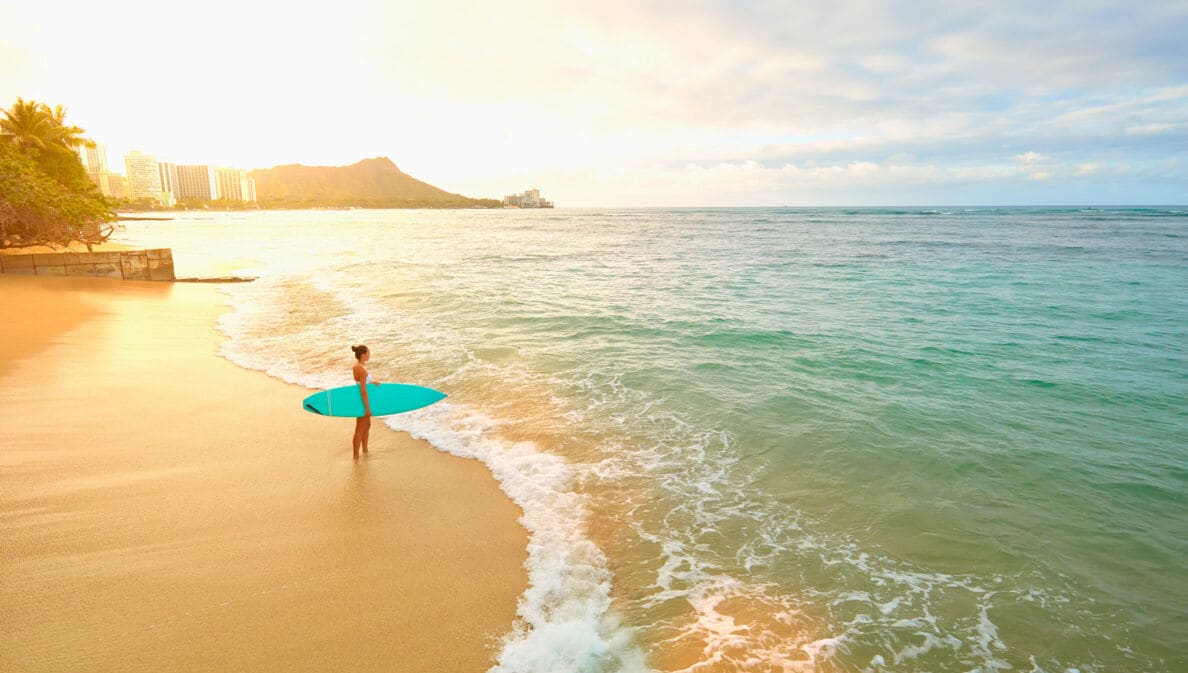 Eine Frau mit türkisfarbenem Surfboard unterm Arm steht am frühen Morgen am leeren Strand von Waikiki.