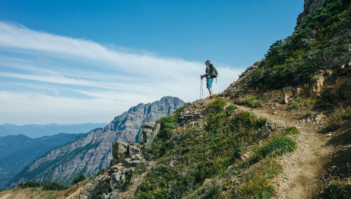 Wanderer mit Rucksack und Wanderstöcken steht auf einem Bergpfad und blickt auf eine Berglandschaft unter blauem Himmel