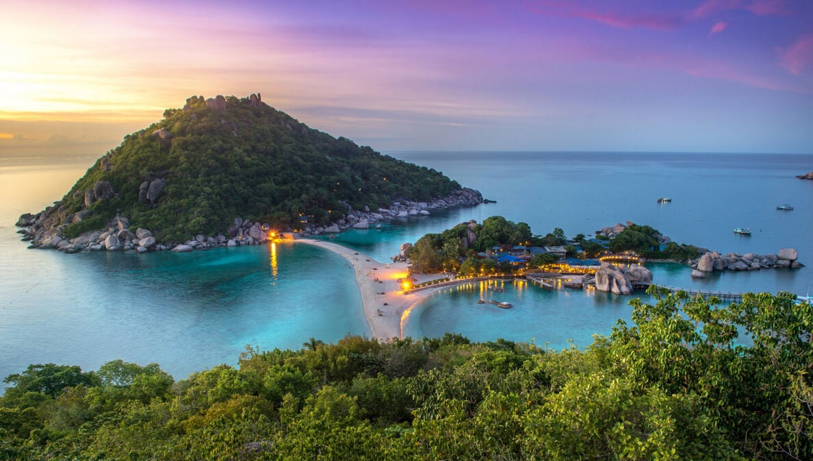 Blick auf eine tropische Insel mit bewaldetem Hügel, Sandbank und beleuchteten Gebäuden bei Sonnenuntergang