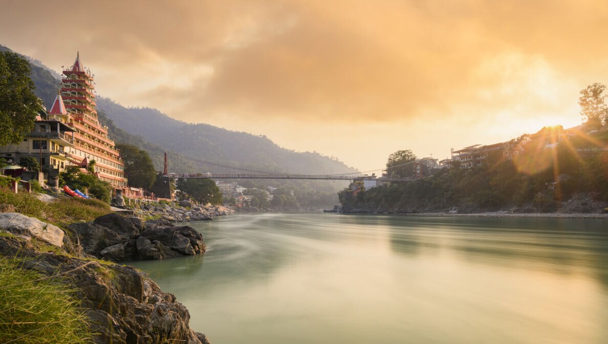 Fluss mit hängender Fußgängerbrücke und mehrstöckigem Tempelgebäude am Ufer bei Sonnenuntergang in bergiger Landschaft